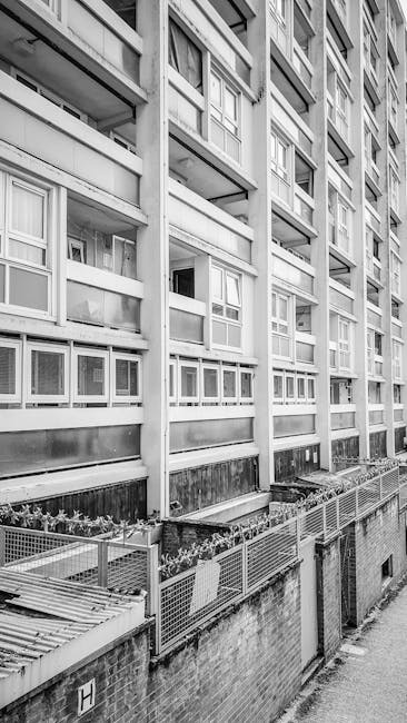 A close-up view of a multi-storey residential apartment building with a concrete facade, featuring multiple rows of windows and balconies. Some balconies are partially enclosed with glass or fabric, w