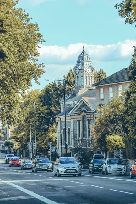 A high-angle view of a busy urban street in Kingston, showing a mix of historic and modern buildings lining both sides of the road. The street features parked cars along the curb, traffic lights, and